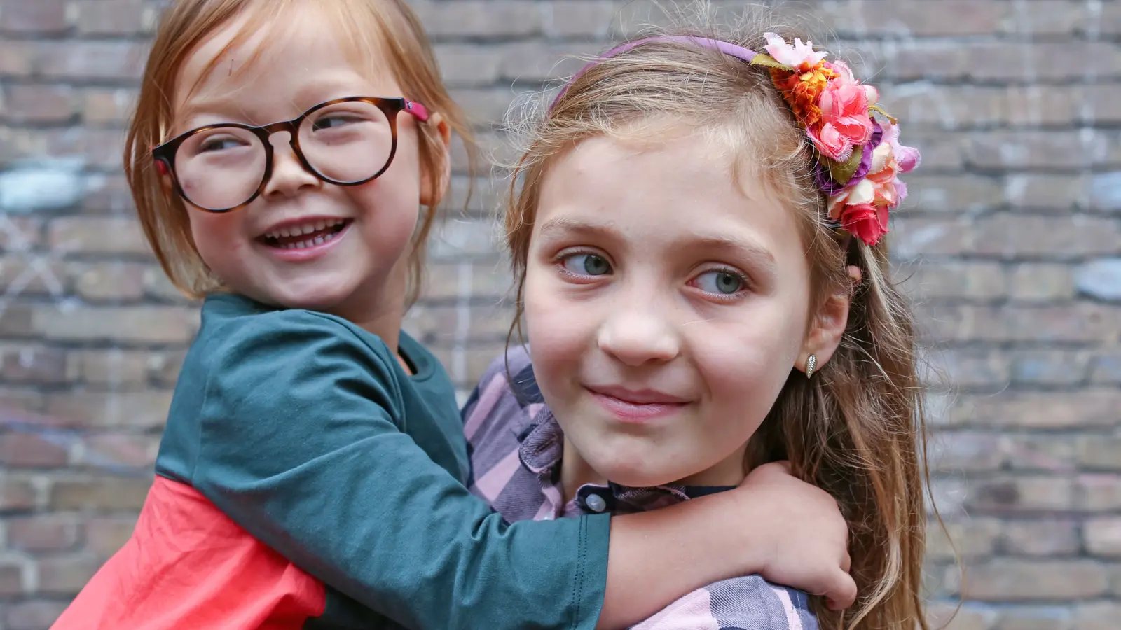 Fotografia przedstawia zbliżenie na dwie uśmiechnięte dziewczynki. Młodsza, po lewej, ma jasne włosy i okulary w brązowych oprawkach, z różowym akcentem na zausznikach. Patrzy prosto w obiektyw z szerokim uśmiechem. Starsza dziewczynka, po prawej, ma na głowie opaskę z kolorowymi kwiatami i również się uśmiecha. W tle widać fragment ceglanego budynku.