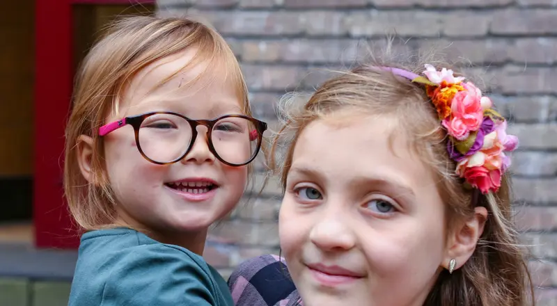 Fotografia przedstawia zbliżenie na dwie uśmiechnięte dziewczynki. Młodsza, po lewej, ma jasne włosy i okulary w brązowych oprawkach, z różowym akcentem na zausznikach. Patrzy prosto w obiektyw z szerokim uśmiechem. Starsza dziewczynka, po prawej, ma na głowie opaskę z kolorowymi kwiatami i również się uśmiecha. W tle widać fragment ceglanego budynku.