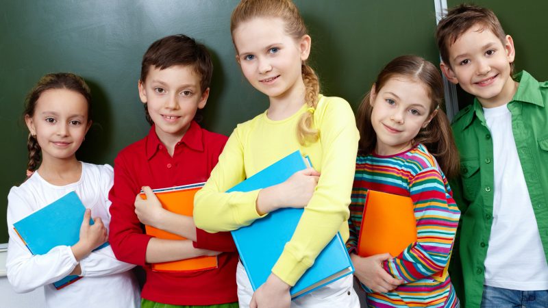 Obrazek przedstawia grupę pięciorga dzieci stojących razem na tle tablicy szkolnej. Każde z nich trzyma w rękach kolorowe zeszyty lub teczki. Dzieci uśmiechają się i wyglądają na zadowolone.