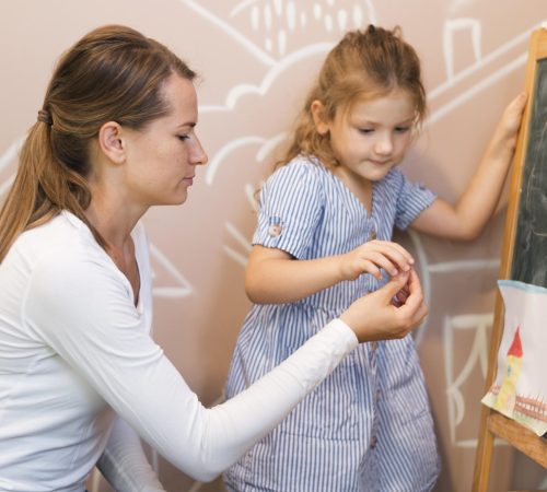 Programy profilaktyczne w przedszkolu - dziewczynka rysująca kredą po tablicy pod okiem przedszkolanki.